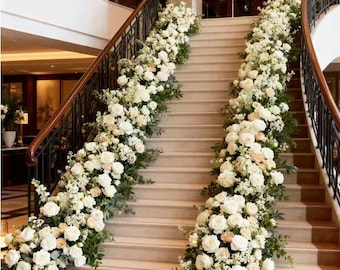 May include: A grand staircase adorned with lush floral arrangements. White roses and other blooms cascade down the steps, accented by greenery. The staircase features marble steps, dark metal railings, and a wooden handrail, creating an elegant ambiance.