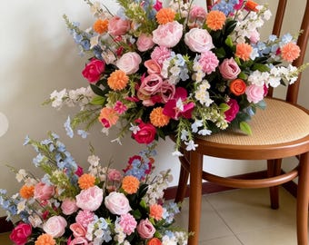 May include: Two floral arrangements with a variety of colorful flowers, including pink roses, orange pom-poms, and blue delphiniums. One arrangement sits on a wooden chair with a woven seat. The flowers are arranged in a lush, full style.
