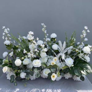 Puede incluir: Arreglo floral rectangular con rosas blancas, hortensias y vegetaci&oacute;n variada. El arreglo incluye hojas grandes de color verde oscuro y delicados acentos blancos, creando una exhibici&oacute;n exuberante y elegante. Ideal para bodas o decoraci&oacute;n del hogar.