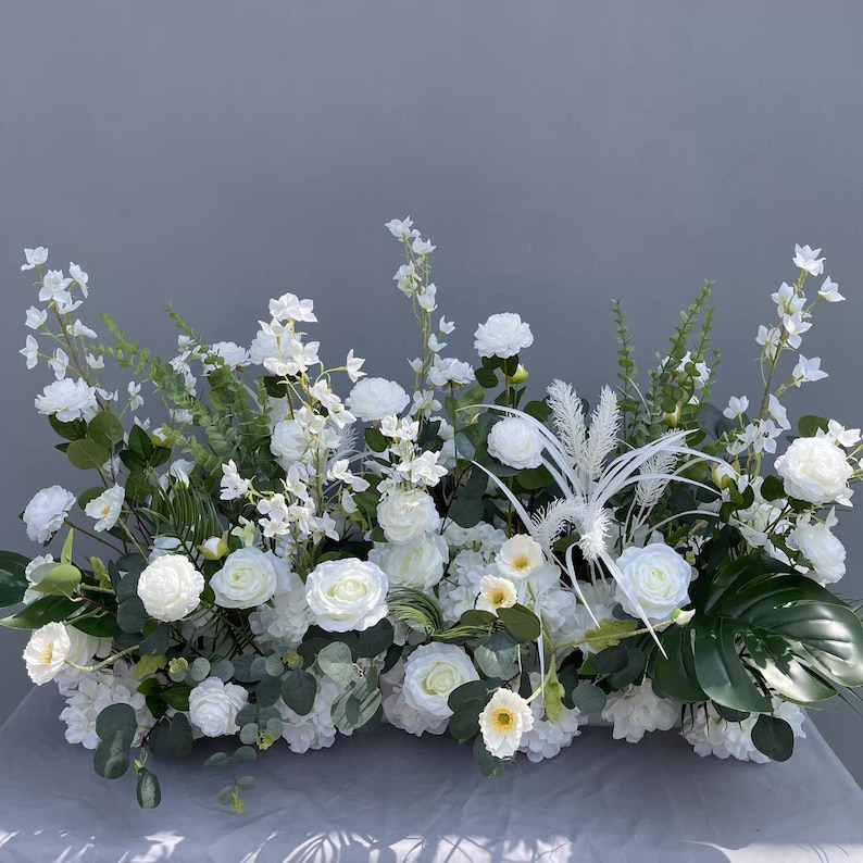 Puede incluir: Arreglo floral rectangular con rosas blancas, hortensias y vegetaci&oacute;n variada. El arreglo incluye hojas grandes de color verde oscuro y delicados acentos blancos, creando una exhibici&oacute;n exuberante y elegante. Ideal para bodas o decoraci&oacute;n del hogar.