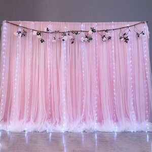 Op de afbeelding: Een roze tule achtergrond met witte sprookjeslichtjes die eroverheen zijn gespannen. De achtergrond is versierd met een tak met kunstbloemen.