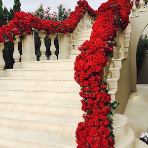 Puede incluir: Una gran escalera exterior adornada con vibrantes rosas rojas. Las rosas caen en cascada por la balaustrada y a lo largo de los escalones, creando una exhibición romántica y elegante. Las escaleras son de piedra clara.