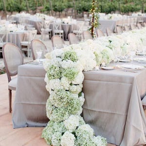 Pode incluir: Uma longa mesa montada para um evento ao ar livre, coberta com uma toalha de mesa cinza e adornada com um arranjo floral em cascata de hortênsias brancas e verdes. A configuração inclui cadeiras, talheres e um vinhedo ao fundo.