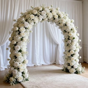 May include: A white floral archway, perfect for weddings or events. The arch is densely covered in white roses, hydrangeas, and baby's breath, creating a full, elegant look. The arch stands on a neutral-colored rug, with white curtains in the background.