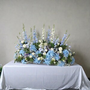 May include: A large floral arrangement with blue hydrangeas, white roses, and blue and white larkspur. The flowers are arranged on a white tablecloth, creating a fresh and elegant display.