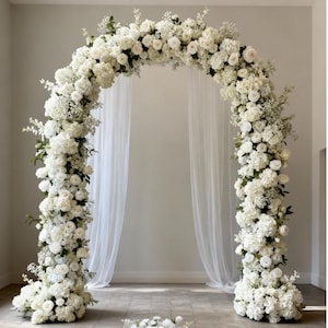 Op de afbeelding: Een witte bloemenboog, versierd met witte rozen, hortensia's en delicate groene bladeren. De boog staat tegen een achtergrond van transparante witte gordijnen, wat een elegante en romantische setting creëert voor een bruiloft of evenement.