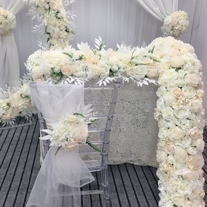 Può includere: Uno sfondo per cerimonia nuziale bianco con un tavolo e sedie bianche decorate con fiori e verde bianco. Il tavolo è coperto da una tovaglia di pizzo bianco e le sedie sono decorate con tulle e fiori bianchi.