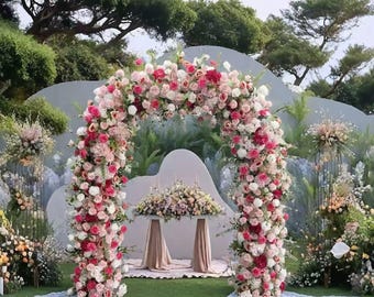 Puede incluir: Un arco floral, adornado con rosas rosas, rojas y blancas, crea un telón de fondo para una ceremonia de boda. El arco está situado sobre un césped verde, con una mesa y arreglos florales. La escena es al aire libre con árboles al fondo.