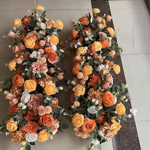 May include: Two floral arrangements with orange, peach, and white roses, carnations, and other flowers. Green foliage complements the blooms. Ideal for weddings or events, these arrangements are visually striking.