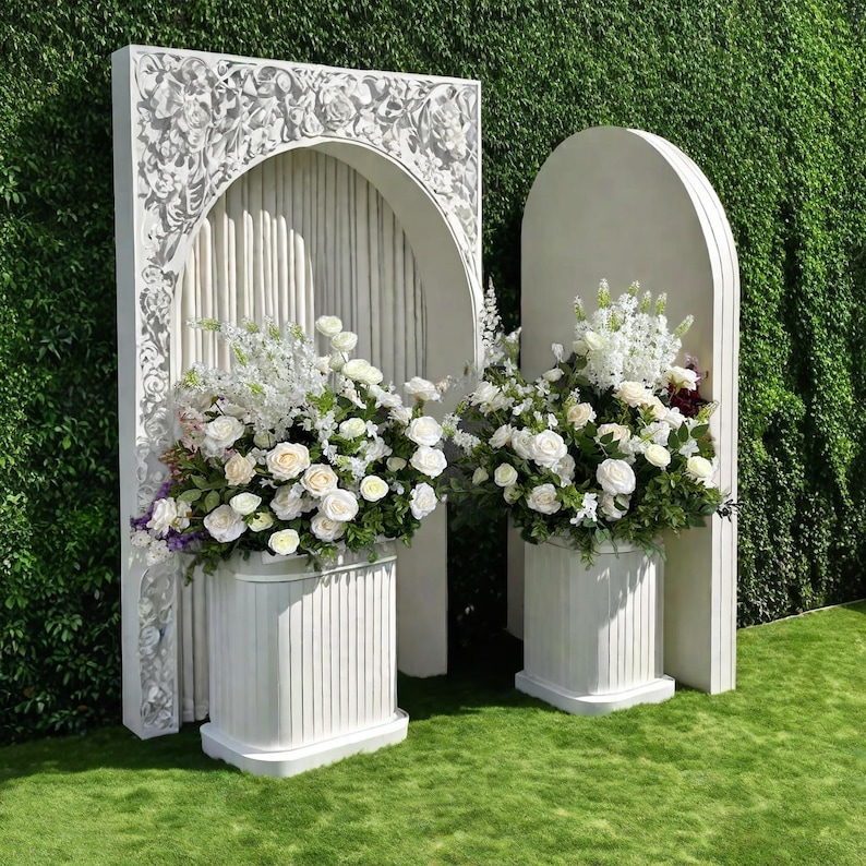May include: Two white decorative structures with floral arrangements. The structures have an arched design and are placed on a green lawn. The floral arrangements feature white roses and other white flowers. The background is a green hedge.
