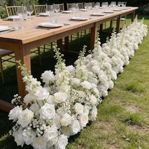 Puede incluir: Una larga mesa de madera preparada para un evento formal, con platos blancos, vasos y cubiertos. Un exuberante arreglo de rosas blancas y otras flores decora la base de la mesa, creando una exhibición floral. La mesa está sobre hierba verde.