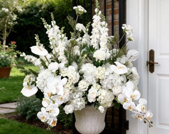 Puede incluir: Una gran urna de piedra ornamentada rebosante de un exuberante arreglo de flores blancas. El arreglo incluye orquídeas, rosas y otras flores, creando una exhibición completa y elegante. La urna se encuentra sobre una base cuadrada cerca de una puerta blanca y una verja de hierro forjado negro.