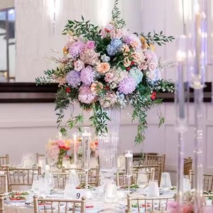 Puede incluir: Un jarrón alto de vidrio transparente lleno de un gran arreglo de flores de color rosa pastel, morado y azul. Las flores están rodeadas de vegetación y están dispuestas en cascada. El jarrón está sentado sobre un mantel blanco en una mesa con sillas doradas.