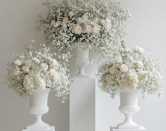 May include: Three white urns filled with white flowers, including roses and baby's breath, are displayed on white pedestals. The arrangements are set against a plain white background, creating a clean and elegant aesthetic. Ideal for weddings or events.