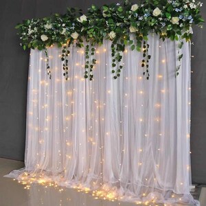 Op de afbeelding: Witte, doorschijnende gordijn achtergrond met witte lichtslingers eroverheen. Een krans van groene bladeren en witte bloemen hangt boven het gordijn.