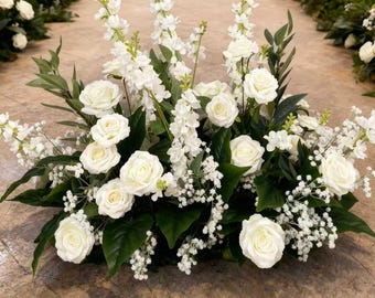 Puede incluir: Arreglo floral con rosas blancas, bocas de dragón y paniculata, con follaje verde. El arreglo está colocado sobre un suelo de piedra, con un pasillo de boda al fondo.