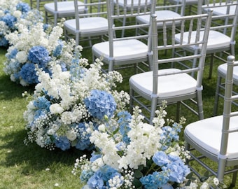 Puede incluir: Montaje para una ceremonia de boda al aire libre con filas de sillas plateadas con cojines blancos. Arreglos florales de hortensias azules y flores blancas bordean el pasillo sobre un césped verde. La escena es brillante y soleada.