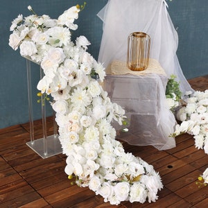 May include: White artificial flower garland featuring orchids, roses, and hydrangeas. The garland is draped over a clear acrylic stand and is ideal for wedding decor or a special event.