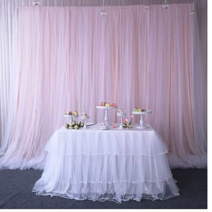 Op de afbeelding: Een witte tule tafelrok met drie lagen, gedrapeerd over een rechthoekige tafel. De tafel is versierd met vijf witte taartstandaards, elk met kleine taarten en gebak. De tafel staat tegen een achtergrond van een roze tule gordijn.