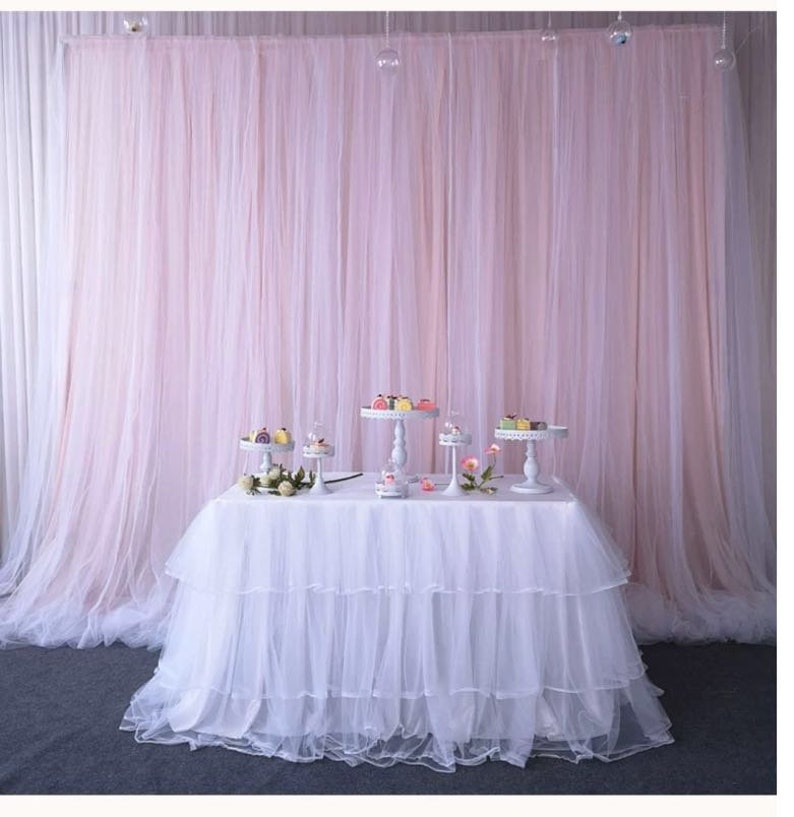 Op de afbeelding: Een witte tule tafelrok met drie lagen, gedrapeerd over een rechthoekige tafel. De tafel is versierd met vijf witte taartstandaards, elk met kleine taarten en gebak. De tafel staat tegen een achtergrond van een roze tule gordijn.