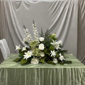 May include: A floral centrepiece arrangement with white lilies, roses, and hydrangeas, complemented by green foliage. The arrangement is displayed on a light green tablecloth, creating a fresh and elegant display.