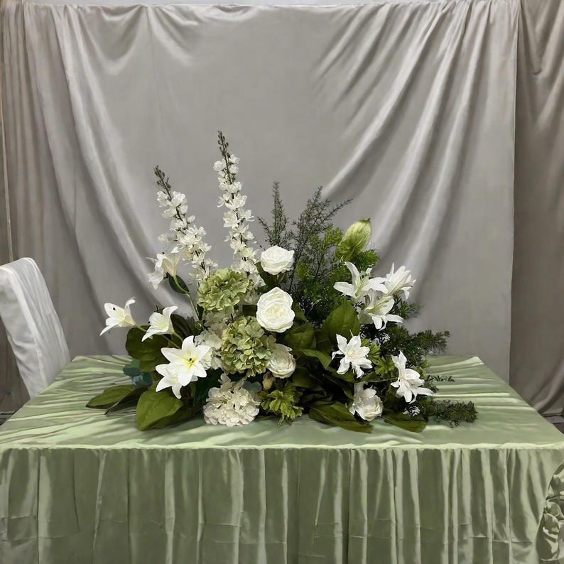 May include: A floral centrepiece arrangement with white lilies, roses, and hydrangeas, complemented by green foliage. The arrangement is displayed on a light green tablecloth, creating a fresh and elegant display.
