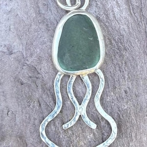 May include: A silver pendant with a sea glass center in a teardrop shape, featuring four wavy tendrils. The pendant has a loop at the top for a chain. The sea glass is a muted green color.