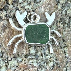 May include: A silver crab pendant with a rectangular, dark green sea glass center. The crab has detailed legs and claws, and a loop for a chain. The pendant is set against a speckled, light brown and gray surface.