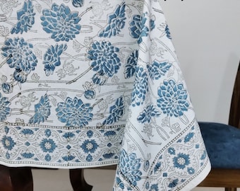 Puede incluir: Un mantel blanco con un patrón floral azul. El mantel tiene un borde de diseños florales azules y blancos. El mantel está drapeado sobre una mesa y una silla de madera.