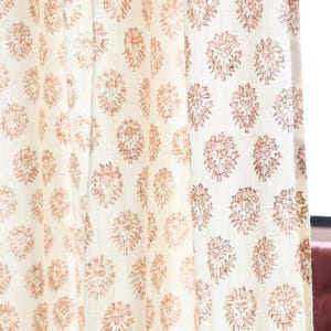 May include: Cream-coloured curtain panels featuring a repeating pattern of coral-coloured floral designs. The fabric has a subtle grid pattern, and the curtains are slightly draped, showing their texture and flow. The overall aesthetic is soft and inviting.
