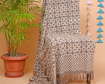 May include: A decorative throw blanket draped over a wooden chair. The blanket features a repeating white floral pattern on a gray background. The bottom edge has a geometric design and fringe.