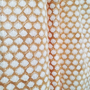May include: A close-up of a fabric featuring a repeating pattern of white seashells against a tan backdrop. The seashells are uniformly arranged, giving a textured look. The fabric is folded, revealing depth and dimension.
