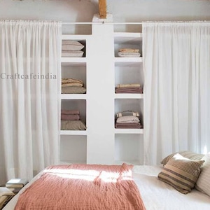 Op de afbeelding: Een witte slaapkamer met een bed met een koraalkleurig dekbed. Het bed staat voor een witte plank met planken vol gevouwen dekens en kussens. Witte, doorschijnende gordijnen hangen aan beide kanten van de plank.