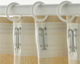 Pode incluir: Varão de cortina branco com três argolas e cortinas bege com uma fita branca e ganchos prateados.