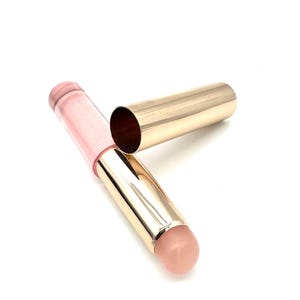 May include: A cylindrical lip balm tube with a metallic gold base and cap, featuring a pale pink translucent upper section and a light peach-colored balm tip exposed at the end. The cap is detached and positioned next to the main tube body.
