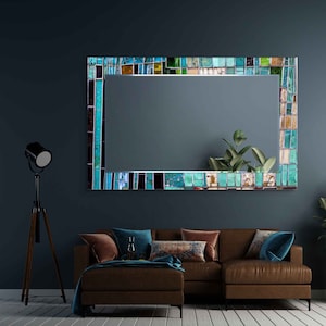 Op de afbeelding: Een grote rechthoekige spiegel met een mozaïeklijst gemaakt van kleine, kleurrijke glazen tegels in tinten blauw, groen en bruin. De spiegel hangt aan een donkerblauwe muur boven een bruine leren bank.