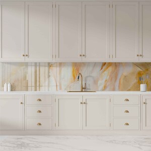 May include: A white kitchen with white cabinets and a marble countertop. The backsplash is a colorful abstract design with gold accents. A gold faucet is mounted above a stainless steel sink.