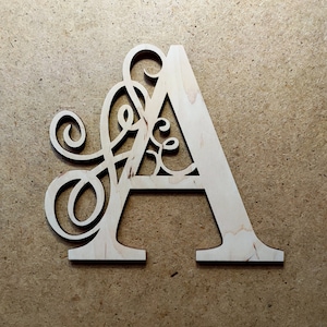 May include: A wooden letter A with a decorative, intricate design cut out of the letter. The letter is light brown and is on a brown background.