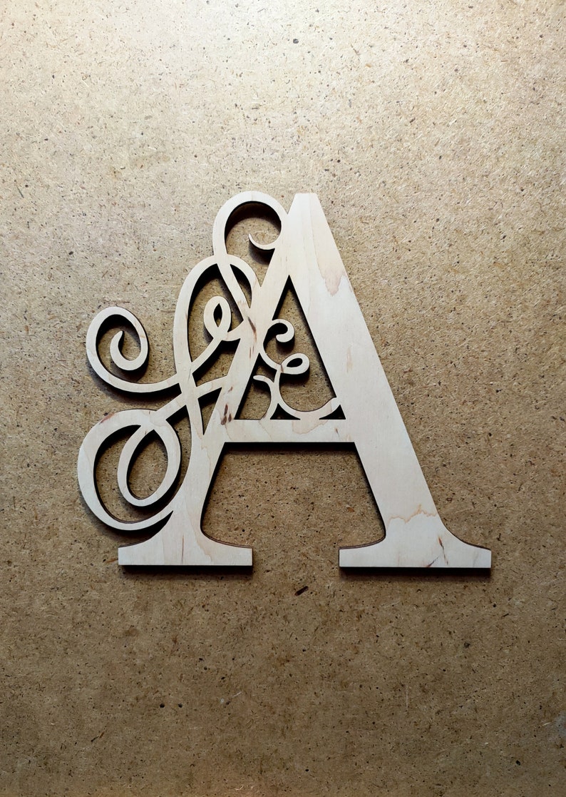 May include: A wooden letter A with a decorative, intricate design cut out of the letter. The letter is light brown and is on a brown background.