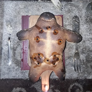 May include: A dark brown, human-like figure with circular markings, placed on a wooden board. Two forks are positioned on either side, and a lit candle is at the base. The background features a patterned rug and a dark sculpture.