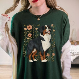 Pu&ograve; includere: Una maglietta verde con un'illustrazione di un cane da montagna bernese circondato da fiori. Il cane &egrave; marrone, nero e bianco con uno sfondo floreale.