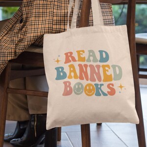 Peut inclure: Un sac en toile blanc avec le texte "READ BANNED BOOKS" en lettres r&eacute;tro color&eacute;es et des smileys. Le sac est accroch&eacute; au dossier d'une chaise en bois.