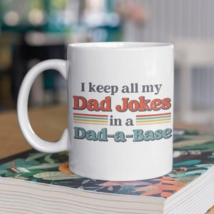 May include: White ceramic mug with a colorful retro design. The mug says "I keep all my Dad Jokes in a Dad-a-Base".