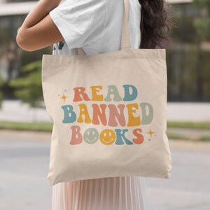 Peut inclure: Une personne portant un sac fourre-tout beige avec le texte "READ BANNED BOOKS" en couleurs arc-en-ciel avec des smileys.
