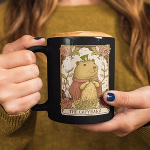 May include: Black ceramic mug with a white illustration of a capybara wearing a crown and a red cape. The illustration is surrounded by a floral border and the text "The Capybara" is printed below the image.