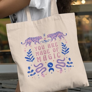 Op de afbeelding: Beige draagtas met een kleurrijk ontwerp. Het ontwerp bevat twee tijgers, de tekst "YOU ARE MADE OF MAGIC", een slang en bloemelementen in roze en blauw. De tas heeft lange beige hengsels.