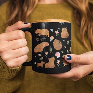 May include: Black ceramic mug with a white design of capybaras, mushrooms, and flowers.