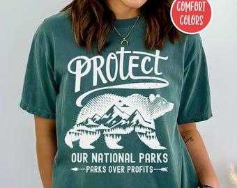 May include: A dark green t-shirt featuring a white graphic of a bear in front of mountains. The text "Protect Our National Parks - Parks Over Profits" is printed on the shirt.