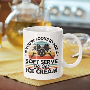 May include: White ceramic mug with a retro-style design featuring two pickleball paddles and a ball. The text reads "If you're looking for a soft serve, go get ice cream."