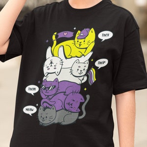 May include: Black t-shirt with a stack of four cartoon cats wearing sunglasses and hats. The cats are stacked on top of each other, with the largest cat at the bottom and the smallest cat at the top. The cats are all different colors, and each cat has a speech bubble with a different pronoun on it. The pronouns are "They", "Their", "Them", and "Meow".